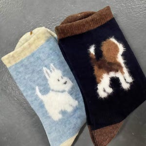 May include: Two pairs of socks with dog designs. One sock is light blue with a white terrier, and the other is navy blue with a brown and white dog. Both socks have brown cuffs and toe sections.