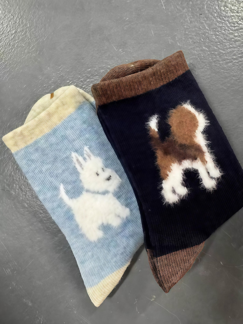 Pu&ograve; includere: Due paia di calzini con disegni di cani. Un calzino &egrave; azzurro con un terrier bianco, e l'altro &egrave; blu navy con un cane marrone e bianco. Entrambi i calzini hanno polsini e punte marroni.