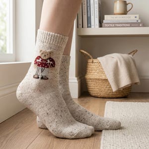 May include: A pair of beige speckled socks with a cartoon bear design. The bear wears a red and white patterned sweater. The socks are worn indoors on a wooden floor, near a window and a shelf with books and a mug.