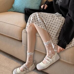 May include: A pair of white and gray Mary Jane style shoes with two straps. The shoes are paired with sheer white socks decorated with small white hearts. The socks have a ruffled trim.