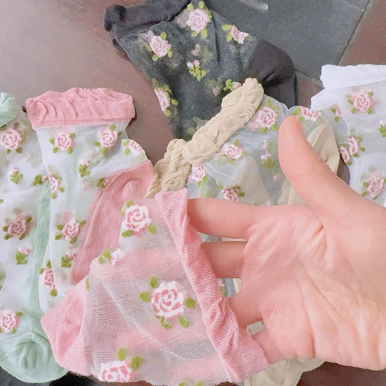 May include: A collection of sheer socks in various colors, including pink, green, and gray. Each sock is adorned with a delicate floral pattern featuring pink roses and green leaves. The socks have a soft, feminine aesthetic.