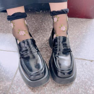 May include: Black platform loafers with a glossy finish. The shoes are paired with sheer black socks embroidered with pink roses. The loafers have a classic penny loafer design with a thick, chunky sole.