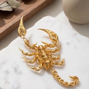 May include: A gold-coloured scorpion pendant with detailed filigree work. The scorpion is displayed on a white marble surface, with a wooden tray and a small vase in the background. The pendant's claws and stinger are clearly visible.
