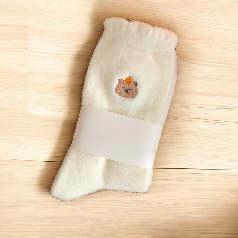 May include: White fluffy socks with a brown bear embroidered on the front. The bear is wearing an orange hat.