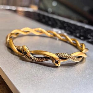 May include: Gold-toned bracelet featuring a snake design. The bracelet has a twisted rope-like pattern with two snake heads meeting at the center. The snakes have textured scales and a star-shaped detail on their heads. The bracelet is on a gray surface.