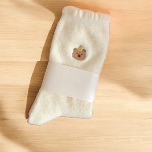 May include: White fluffy socks with a brown bear wearing a yellow crown embroidered on the front. The socks are rolled up and packaged with a white label.