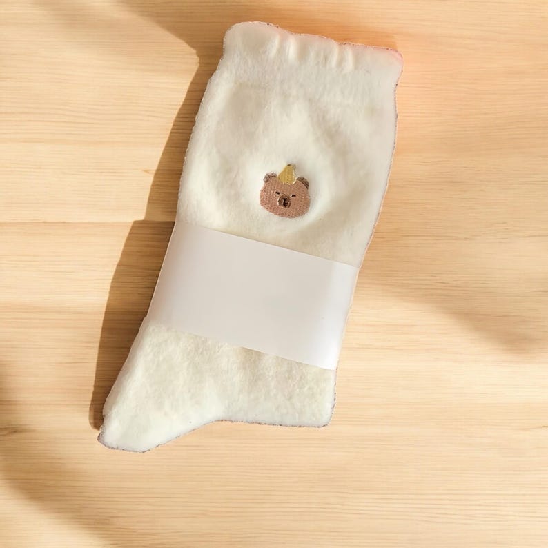 May include: White fluffy socks with a brown bear wearing a yellow crown embroidered on the front. The socks are rolled up and packaged with a white label.