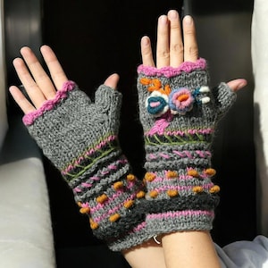 May include: Gray fingerless gloves with pink, orange, and green embroidered details. The gloves have a pink trim and are decorated with colorful floral accents. The design includes raised orange bobbles and intricate stitching.