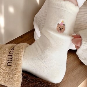 May include: White socks with a brown bear wearing a pink party hat embroidered on the front. The socks are being pulled up on a person's foot.