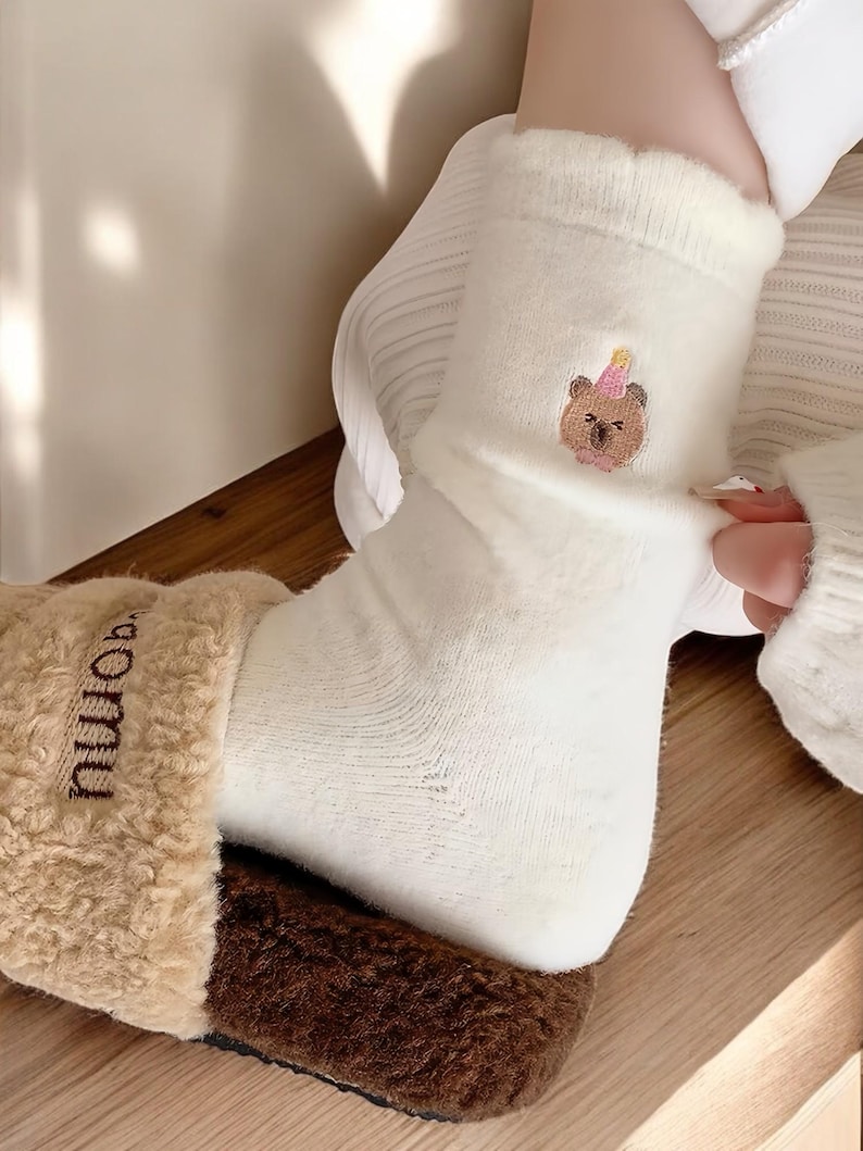 May include: White socks with a brown bear wearing a pink party hat embroidered on the front. The socks are being pulled up on a person's foot.