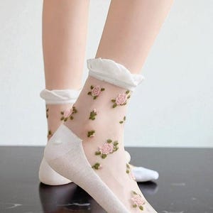 Puede incluir: Calcetines blancos transparentes con bordado de rosas rosas y volantes. Los calcetines presentan un delicado estampado floral. Están hechos de un material ligero y aireado y son visibles en los pies.