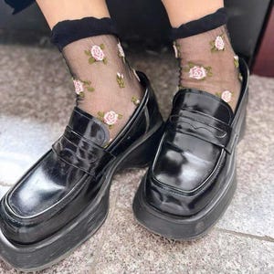 May include: Black platform loafers with a glossy finish are paired with sheer black socks adorned with pink rose patterns. The shoes have a chunky sole and a classic penny loafer design, offering a blend of vintage and modern style.