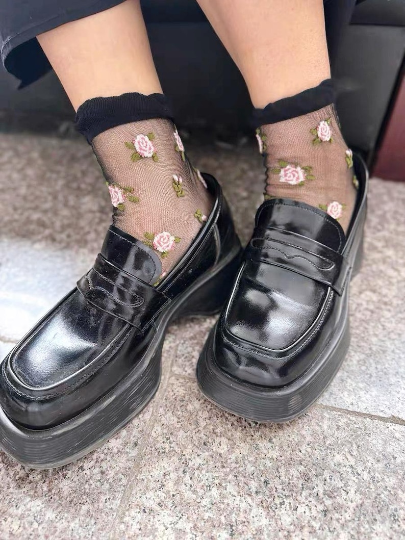 May include: Black platform loafers with a glossy finish are paired with sheer black socks adorned with pink rose patterns. The shoes have a chunky sole and a classic penny loafer design, offering a blend of vintage and modern style.
