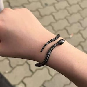 May include: A black snake bracelet coiled around a wrist. The bracelet features a textured, scale-like design and a snake head. The jewellery is made of dark metal and is worn on the arm.