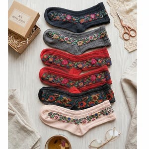 Könnte beinhalten: Eine Auswahl an bestickten Knöchelsocken in Grau-, Rot- und Rosatönen mit floralen Mustern. Die Socken sind auf einer hellen Holzoberfläche zusammen mit einer Schachtel mit der Aufschrift "A's BOHO SOCKS" drapiert.
