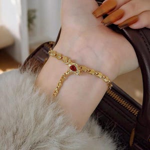 May include: A gold-colored snake bracelet with a red gemstone eye and small clear stones. The bracelet is worn on a wrist, with a matching ring-like band extending to the middle finger.