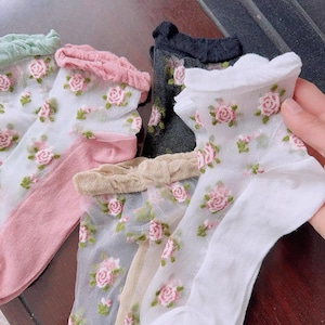 May include: Sheer socks in various colors, including white, pink, and black, adorned with a delicate floral rose pattern. The socks have ruffled edges and are made of a lightweight, see-through material. The roses are pink and green.
