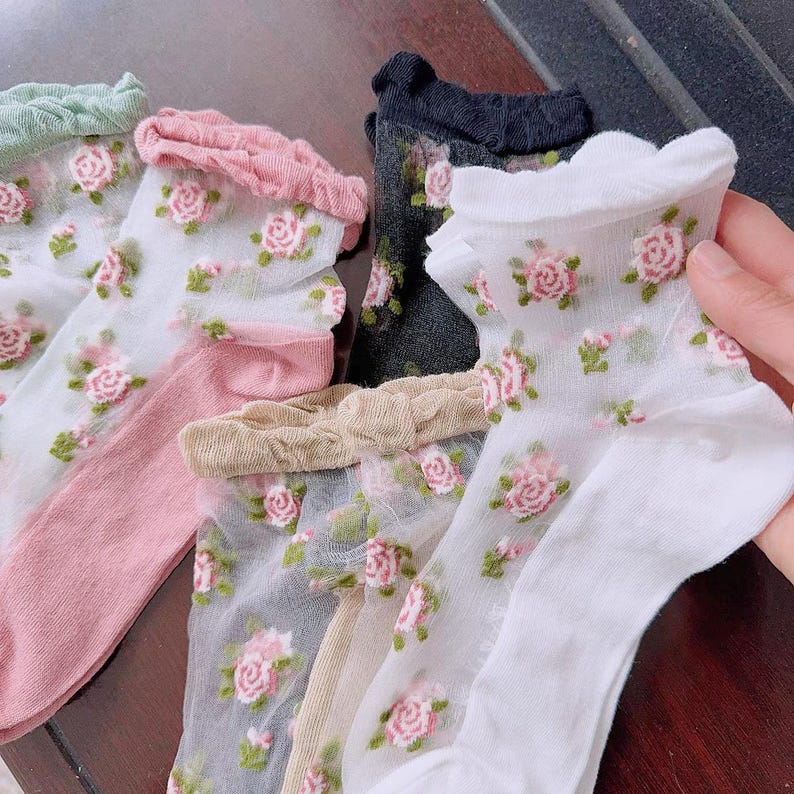 May include: Sheer socks in various colors, including white, pink, and black, adorned with a delicate floral rose pattern. The socks have ruffled edges and are made of a lightweight, see-through material. The roses are pink and green.