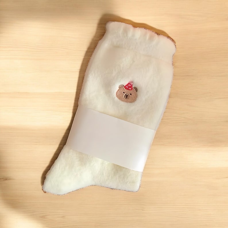 May include: White fluffy socks with a brown bear wearing a red and white party hat embroidered on the front.