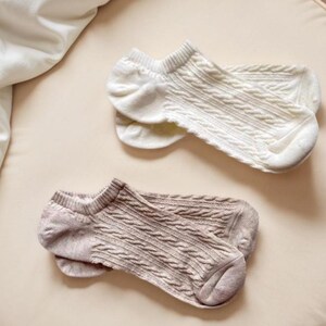 May include: Two pairs of ankle socks in cream and taupe. The socks feature a cable knit design and are made from a soft material. The socks are displayed on a neutral background, suggesting a focus on comfort and style.