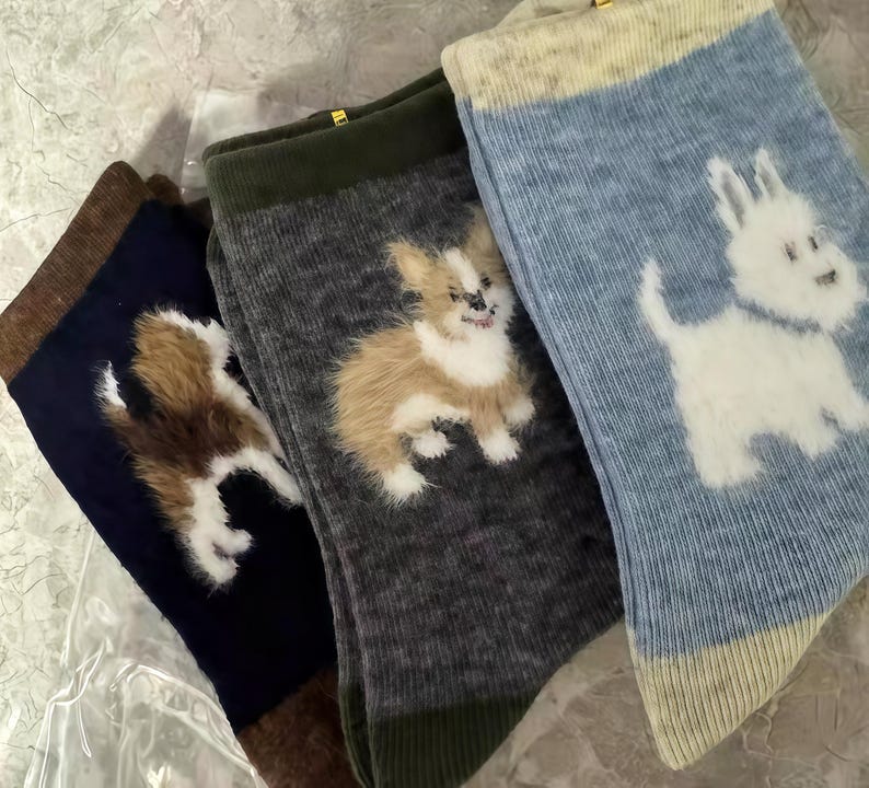 Pu&ograve; includere: Tre paia di calzini con disegni di cani. Un paio &egrave; blu navy con un cane marrone e bianco, un altro &egrave; grigio con un cane marrone chiaro e bianco, e l'ultimo &egrave; azzurro con un cane bianco. I calzini hanno una trama a coste.