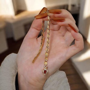 May include: A gold-colored earring with a dangling design. The earring features a series of connected shapes, culminating in a red, heart-shaped stone. The earring is held in a hand.