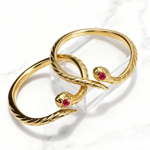 Könnte beinhalten: Zwei goldfarbene Armbänder in Schlangenform. Jedes Armband hat einen Schlangenkopf mit einem roten Edelsteinauge und einen strukturierten Körper. Die Armbänder werden auf einer transparenten Oberfläche vor weißem Hintergrund präsentiert.