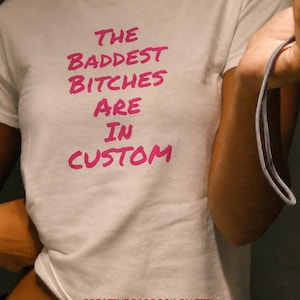 May include: A white short-sleeved t-shirt featuring the text THE BADDEST BITCHES ARE IN CUSTOM printed in a bold, bright pink font across the chest. The shirt has a crew neckline and a standard fit, with the text CREATIVE PADDOCK ON ETSY at the bottom.