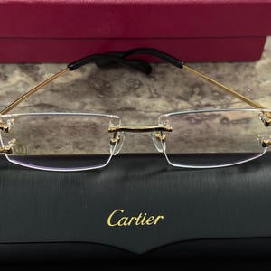 May include: Rimless eyeglasses with gold-tone frames and black temple tips. The glasses rest on a black case with the gold-lettered brand name "Cartier". A burgundy box is visible in the background.
