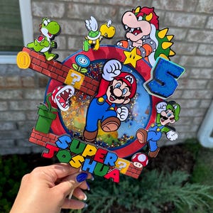 May include: A colorful cake topper featuring Mario, Luigi, Yoshi, Bowser, and a turtle. The topper is decorated with a red, blue, and yellow background and the words "SUPER JOSHUA".
