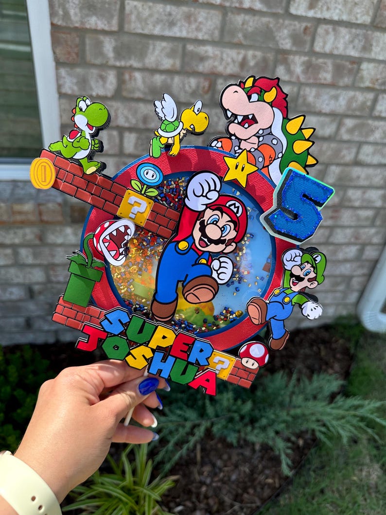 May include: A colorful cake topper featuring Mario, Luigi, Yoshi, Bowser, and a turtle. The topper is decorated with a red, blue, and yellow background and the words "SUPER JOSHUA".