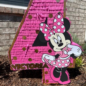 Pu&ograve; includere: Una grande pignatta rosa a forma di numero quattro con un ritaglio di Minnie Mouse. La pignatta &egrave; decorata con frange rosa e dettagli dorati. Il nome "Victoria" &egrave; visualizzato sulla pignatta.