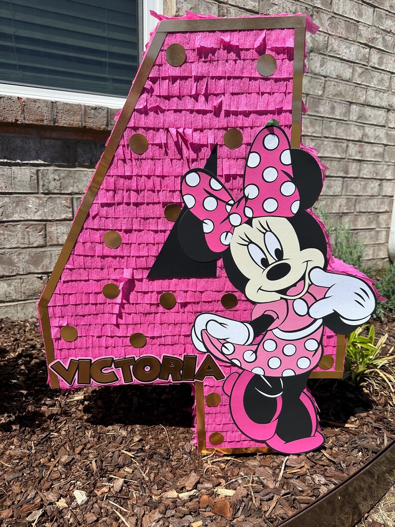 Pu&ograve; includere: Una grande pignatta rosa a forma di numero quattro con un ritaglio di Minnie Mouse. La pignatta &egrave; decorata con frange rosa e dettagli dorati. Il nome "Victoria" &egrave; visualizzato sulla pignatta.