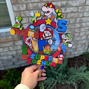 May include: A colorful cake topper featuring Mario, Luigi, Yoshi, and Bowser characters from the Super Mario Bros. video game. The topper includes a red and blue background with a question mark block, a green pipe, and a star. The text "SUPER JOSHUA" is written on the topper.