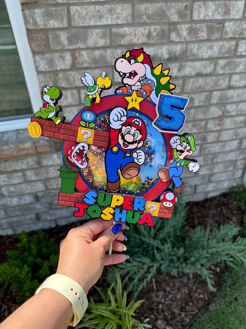 May include: A colorful cake topper featuring Mario, Luigi, Yoshi, and Bowser characters from the Super Mario Bros. video game. The topper includes a red and blue background with a question mark block, a green pipe, and a star. The text "SUPER JOSHUA" is written on the topper.