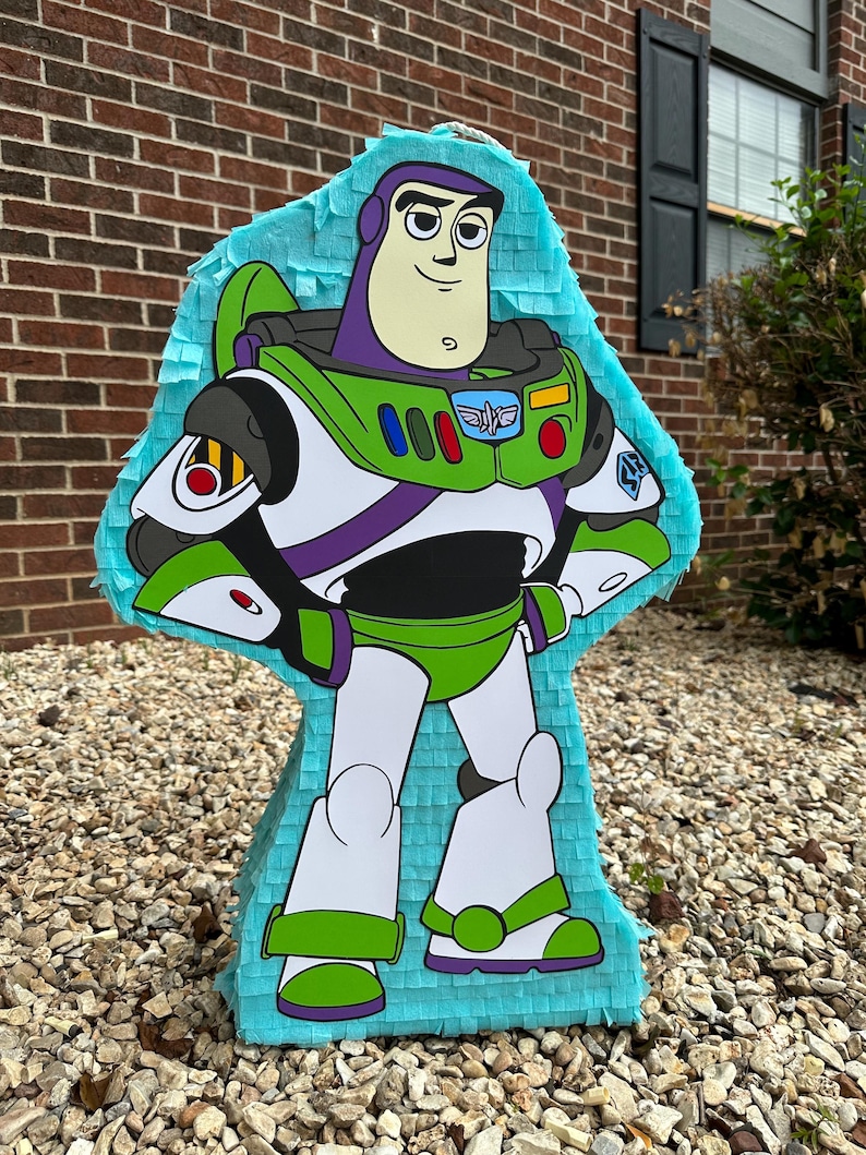 May include: A blue and green Buzz Lightyear pi&ntilde;ata with a white background. The pi&ntilde;ata is made of tissue paper and has a string attached to the top.