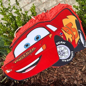 以下が含まれることがあります： 「Rust-eze」と「Lightning McQueen」と書かれた、赤と黄色のカートゥーンカーの形をしたピニャータです。