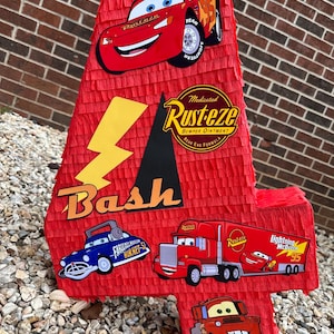 May include: A red number four pi&ntilde;ata decorated with Cars movie characters. The pi&ntilde;ata features Lightning McQueen, Mater, and other characters. The word "Bash" is written on the pi&ntilde;ata, along with the Rust-eze logo.