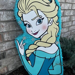 Puede incluir: Una pi&ntilde;ata azul y blanca con forma de Elsa de la pel&iacute;cula Frozen. La pi&ntilde;ata tiene una cara detallada con ojos azules y un rubor rosado. Elsa lleva un vestido azul con un cuello blanco y una trenza en el pelo.