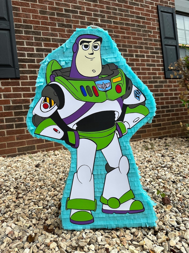 May include: A blue and green Buzz Lightyear pinata. The pinata is shaped like Buzz Lightyear from the Toy Story movies. The pinata has a purple stripe across the chest and a yellow and red stripe across the shoulders. The pinata is standing on a bed of small rocks.