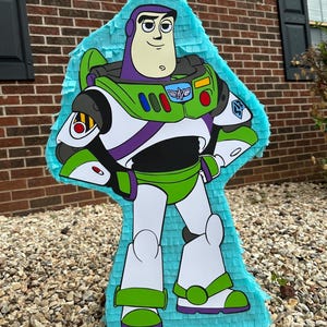 May include: A blue and green Buzz Lightyear pi&ntilde;ata with a white space suit and purple accents. The pi&ntilde;ata is made of tissue paper and has a string hanging from the top.