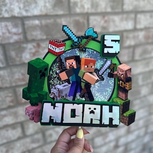 May include: A Minecraft-themed cake topper featuring pixelated characters, weapons, and the name "Noah". The design includes a green Creeper, a pig, a wolf, and the number "5". The background is a green circle with a glittery effect.