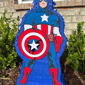 May include: A blue and red Captain America pi&ntilde;ata with a white star on his chest and a red, white and blue shield. The pi&ntilde;ata is made of tissue paper and is ready to be filled with sweets and toys.