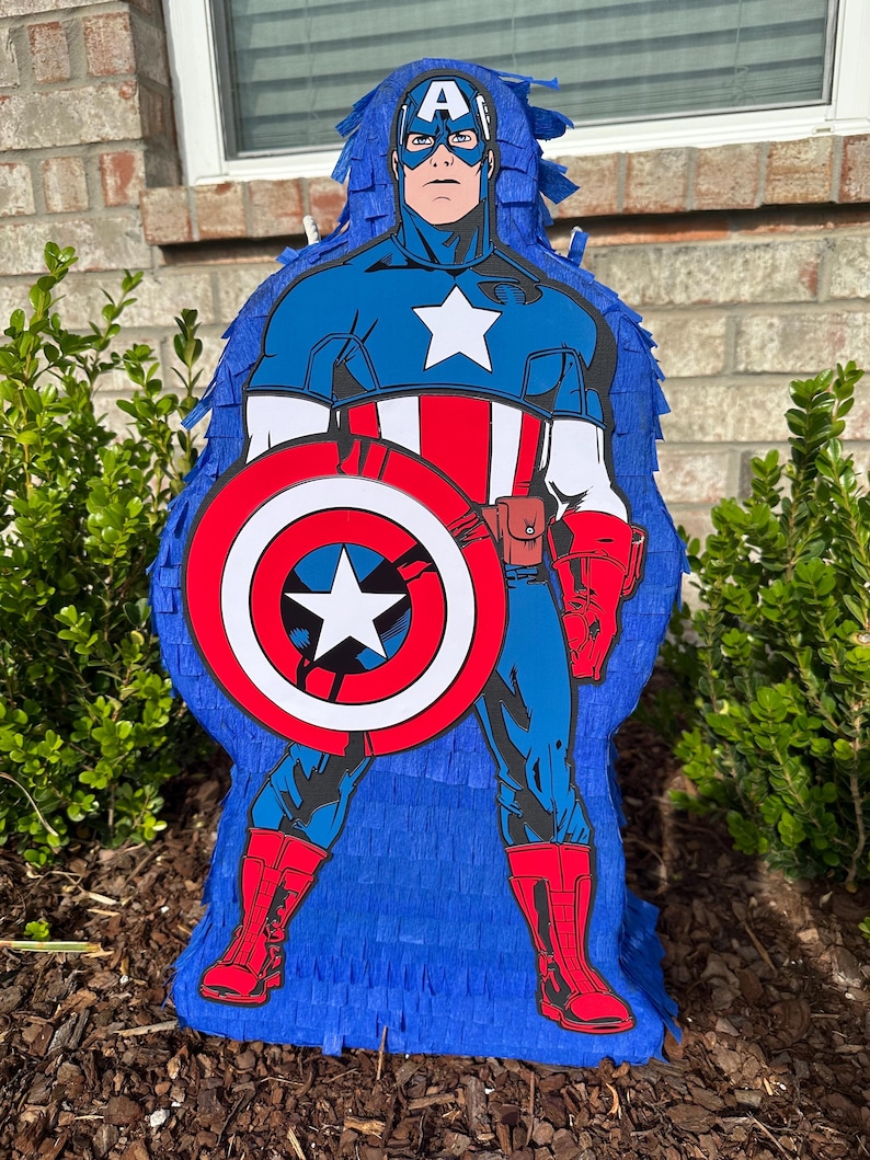 May include: A blue and red Captain America pi&ntilde;ata with a white star on his chest and a red, white and blue shield. The pi&ntilde;ata is made of tissue paper and is ready to be filled with sweets and toys.