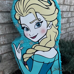 Puede incluir: Una pi&ntilde;ata azul y blanca con forma de Elsa de la pel&iacute;cula Frozen. La pi&ntilde;ata tiene una ilustraci&oacute;n detallada de la cara y el cabello de Elsa. Hay copos de nieve en la pi&ntilde;ata.