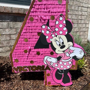Pu&ograve; includere: Una pignatta rosa a forma di numero 4 con dettagli dorati e un ritaglio di Minnie Mouse. La pignatta &egrave; decorata con frange rosa e cerchi dorati. Il nome "Victoria" &egrave; stampato.