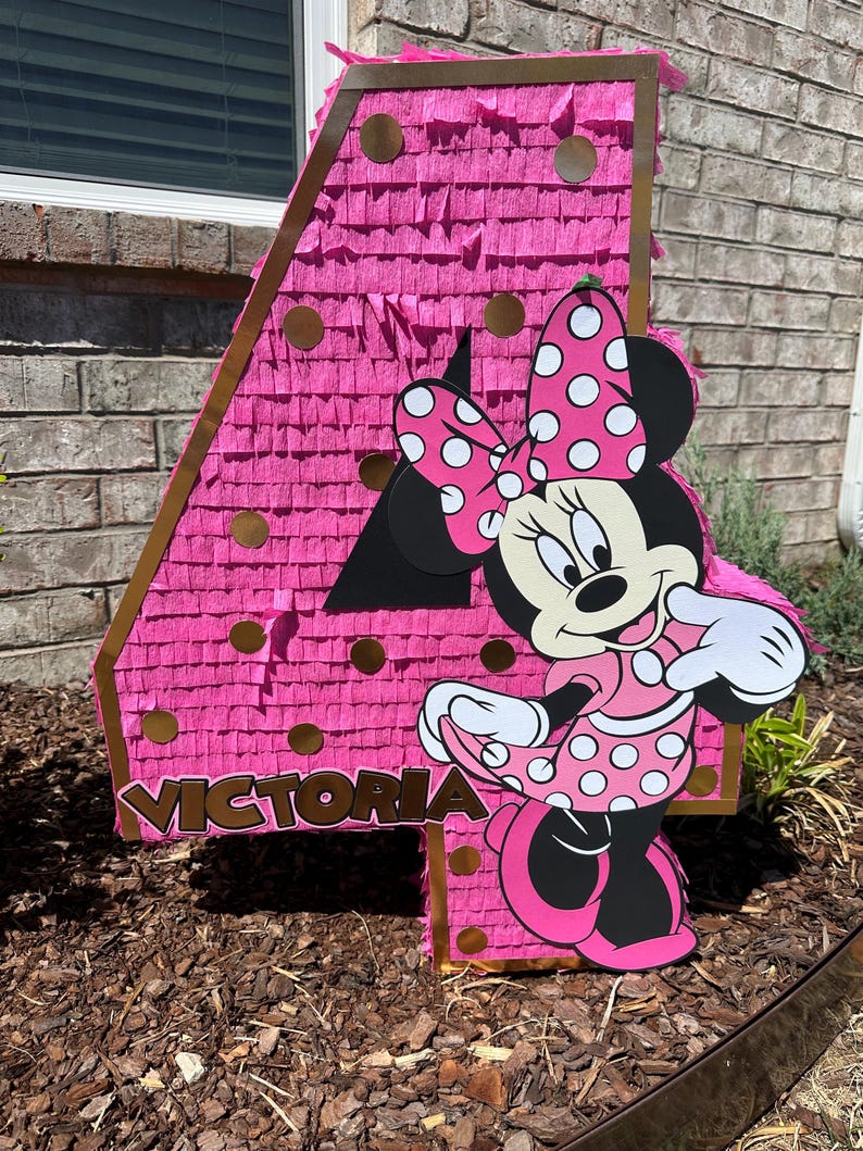 Pu&ograve; includere: Una pignatta rosa a forma di numero 4 con dettagli dorati e un ritaglio di Minnie Mouse. La pignatta &egrave; decorata con frange rosa e cerchi dorati. Il nome "Victoria" &egrave; stampato.