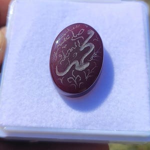 May include: Oval, burgundy-colored gemstone with silver Arabic calligraphy. The stone is set in a white plastic case. The calligraphy appears to be a religious inscription. The gemstone is smooth and polished, reflecting light.