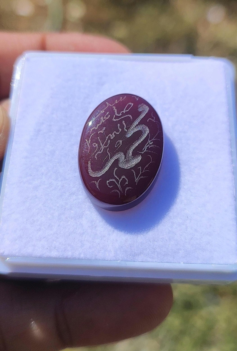May include: Oval, burgundy-colored gemstone with silver Arabic calligraphy. The stone is set in a white plastic case. The calligraphy appears to be a religious inscription. The gemstone is smooth and polished, reflecting light.