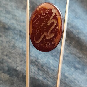 May include: A polished, oval-shaped, reddish-brown gemstone held by tweezers. The stone features an etched, swirling design in a lighter color, possibly depicting calligraphy or a stylized pattern. The background is a blue fabric.